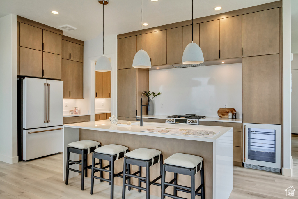 Kitchen with high end fridge, decorative light fixtures, light hardwood / wood-style flooring, wine cooler, and an island with sink