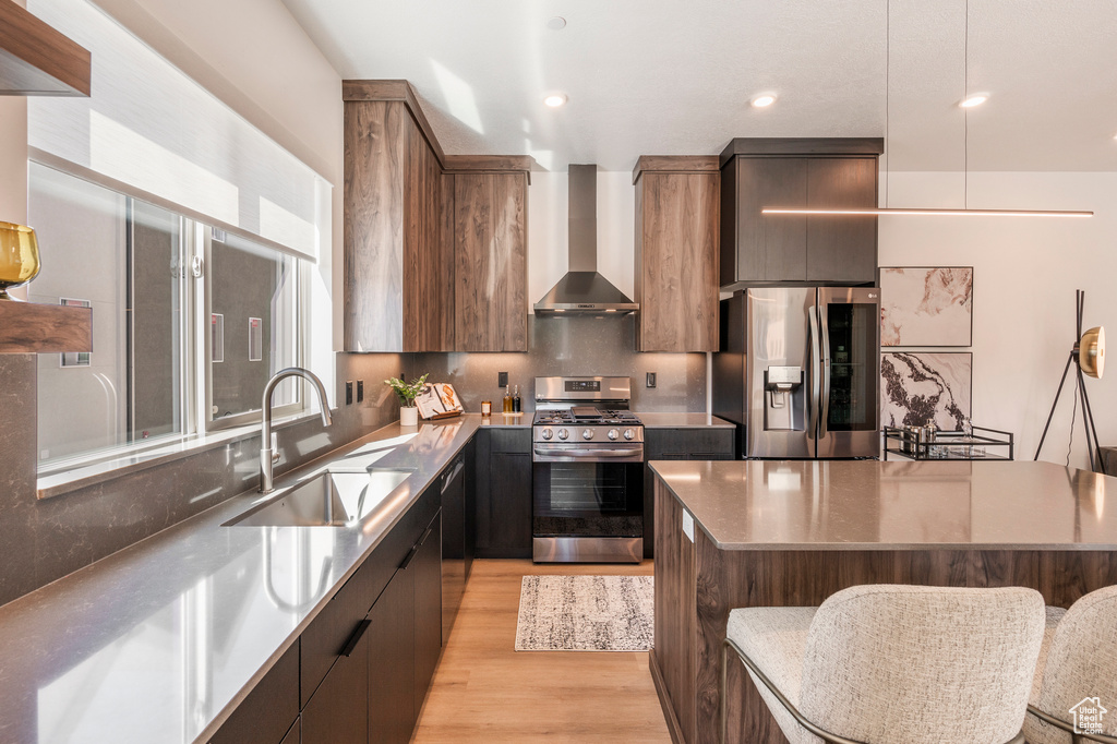 Kitchen featuring stainless steel appliances, light hardwood / wood-style floors, wall chimney exhaust hood, sink, and a center island