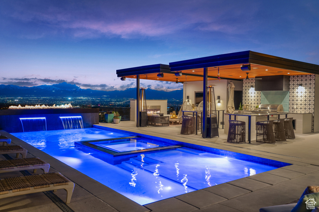 Pool at dusk with pool water feature, a patio area, exterior bar, and an in ground hot tub