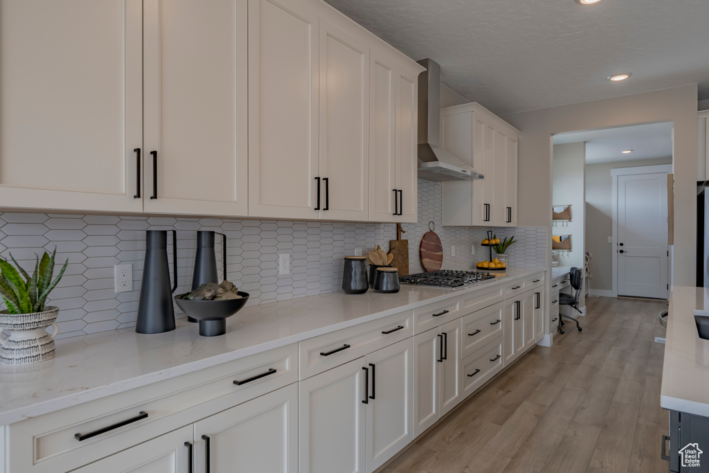 Kitchen with wall chimney range hood, light wood-type flooring, tasteful backsplash, white cabinetry, and light stone countertops