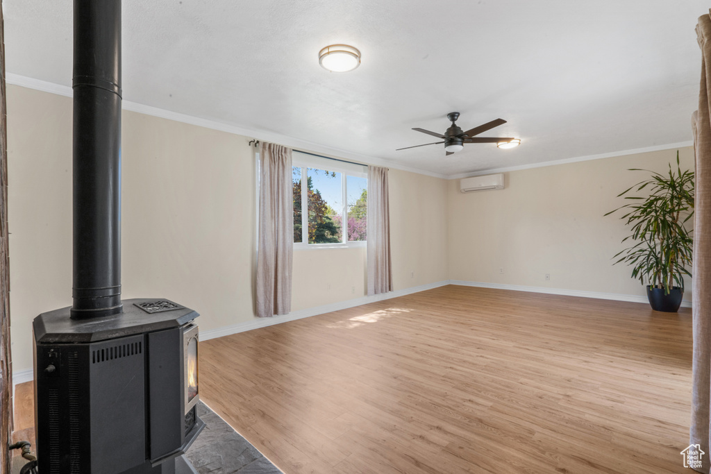 Unfurnished living room featuring ceiling fan, light hardwood / wood-style floors, crown molding, a wall mounted air conditioner, and a wood stove