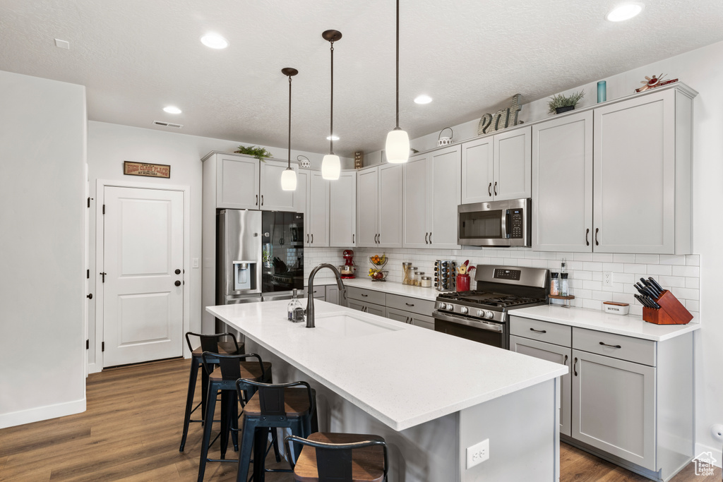 Kitchen with stainless steel appliances, wood-type flooring, tasteful backsplash, an island with sink, and pendant lighting