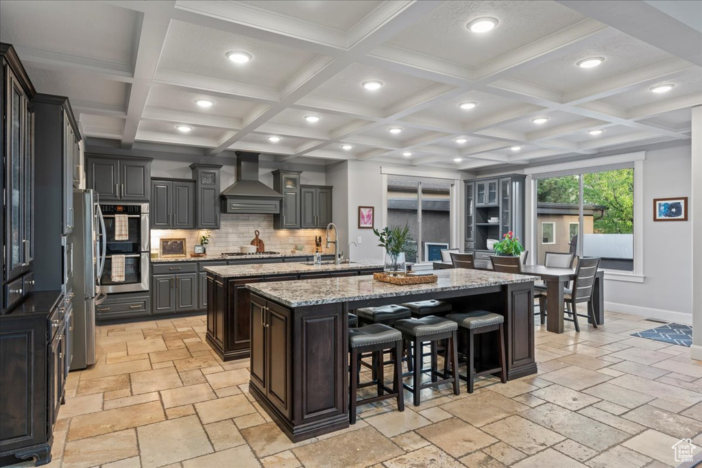 Kitchen with tasteful backsplash, coffered ceiling, wall chimney exhaust hood, and a kitchen island with sink