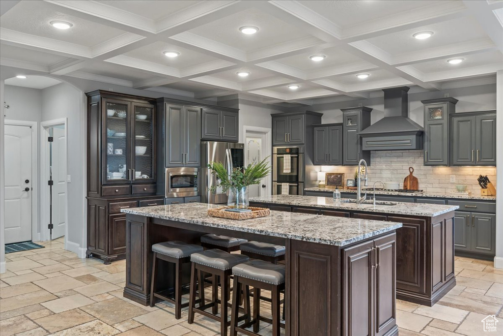 Kitchen with wall chimney range hood, stainless steel appliances, a kitchen island with sink, and coffered ceiling