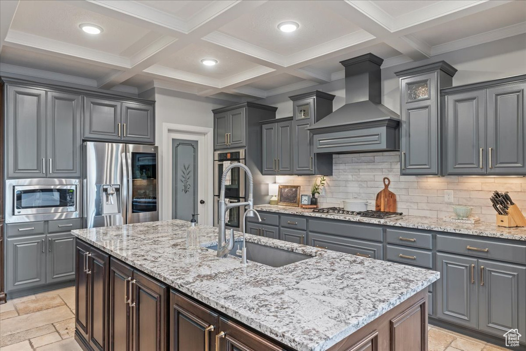 Kitchen featuring appliances with stainless steel finishes, light tile flooring, wall chimney range hood, backsplash, and a center island with sink