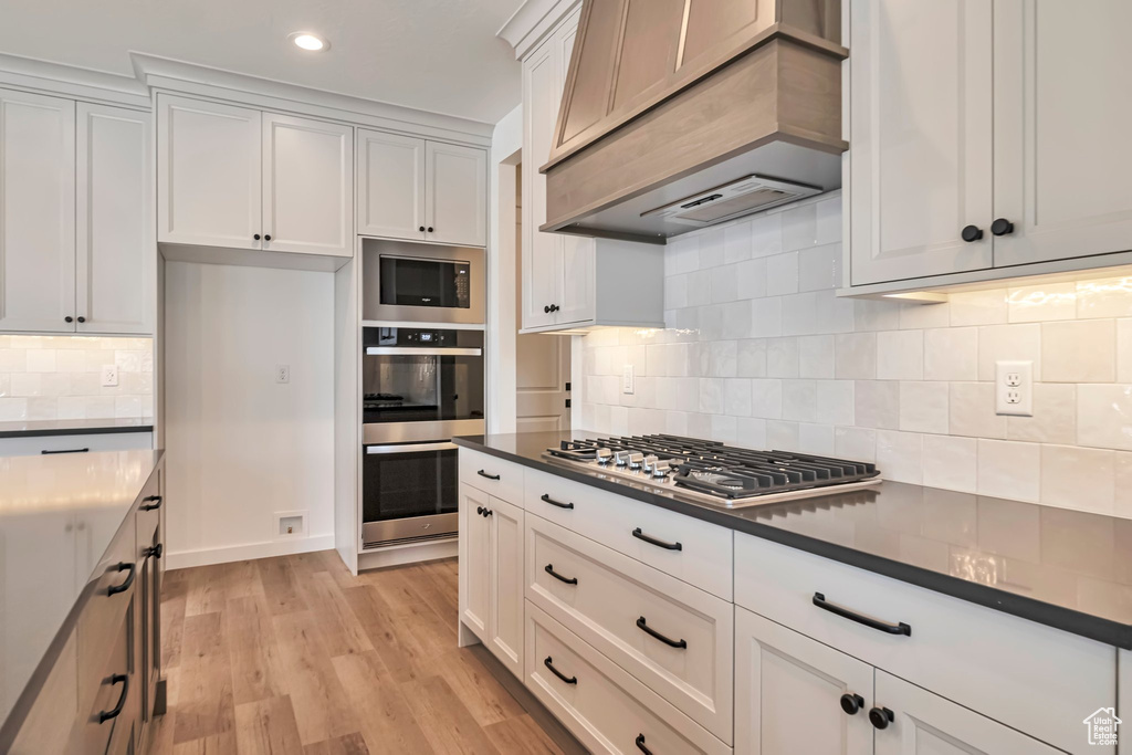 Kitchen with premium range hood, light hardwood / wood-style flooring, white cabinetry, backsplash, and stainless steel appliances