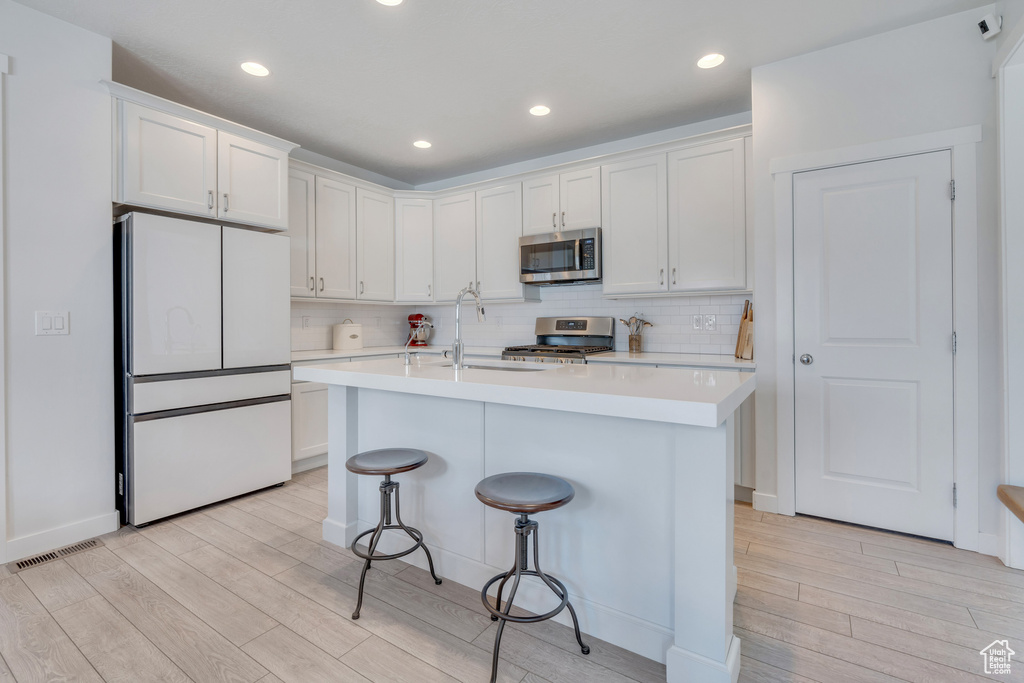 Kitchen featuring white cabinets, backsplash, appliances with stainless steel finishes, a center island with sink, and light hardwood / wood-style floors