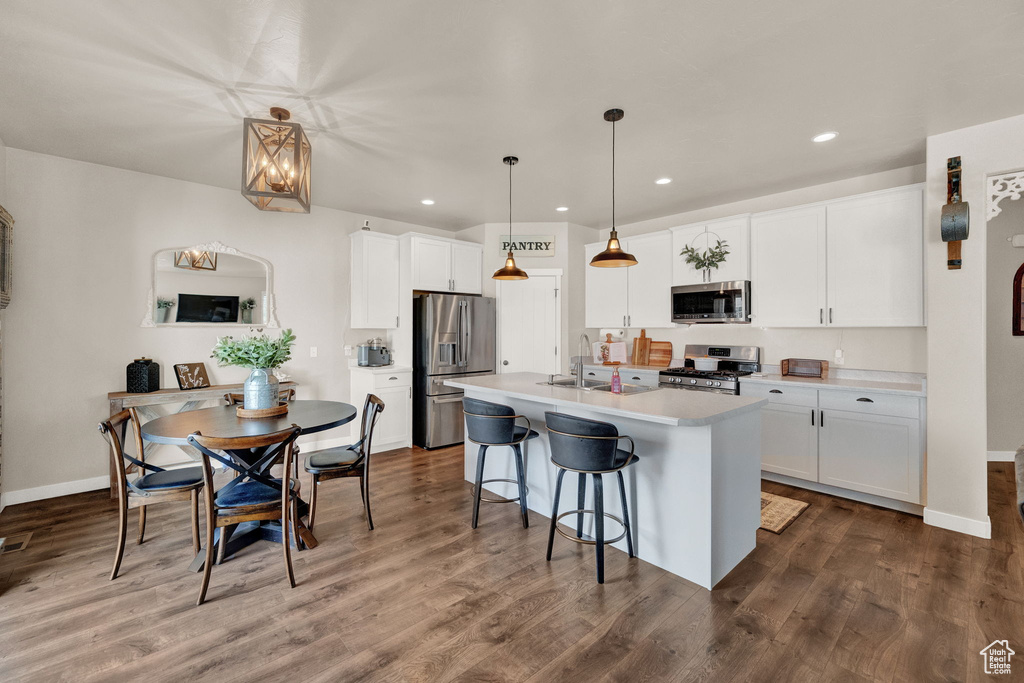 Kitchen with wood-type flooring, decorative light fixtures, white cabinets, appliances with stainless steel finishes, and an island with sink