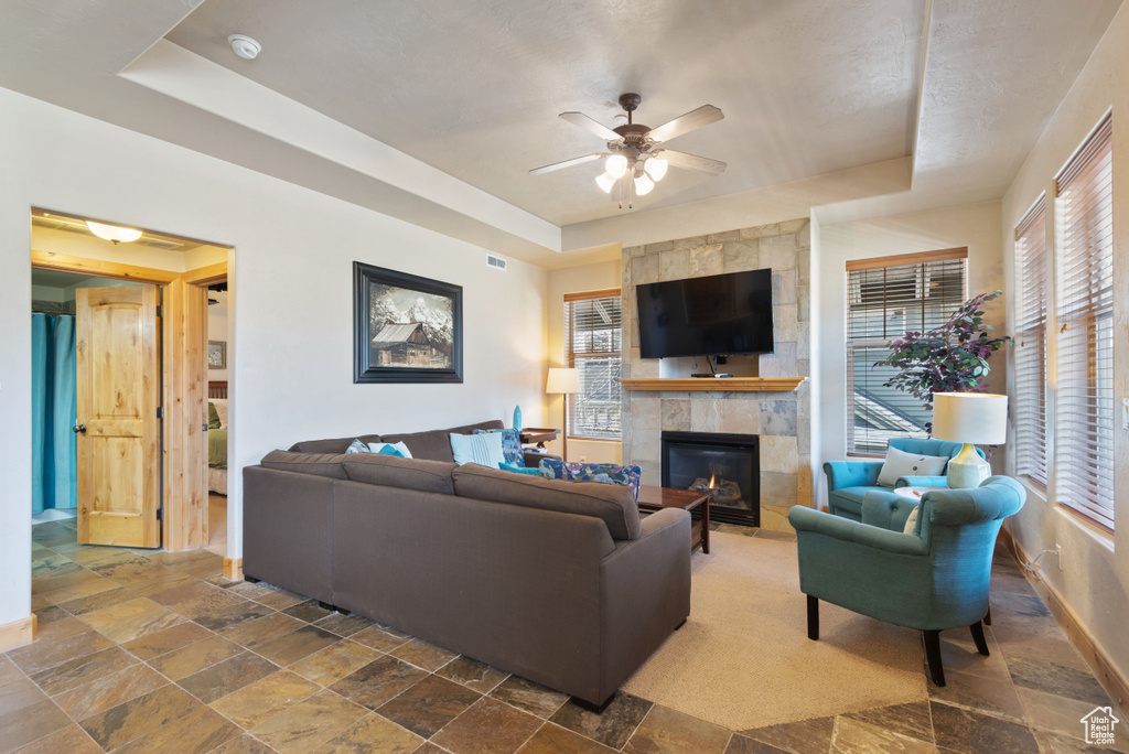 Living room featuring a stone fireplace, tile flooring, ceiling fan, and a tray ceiling
