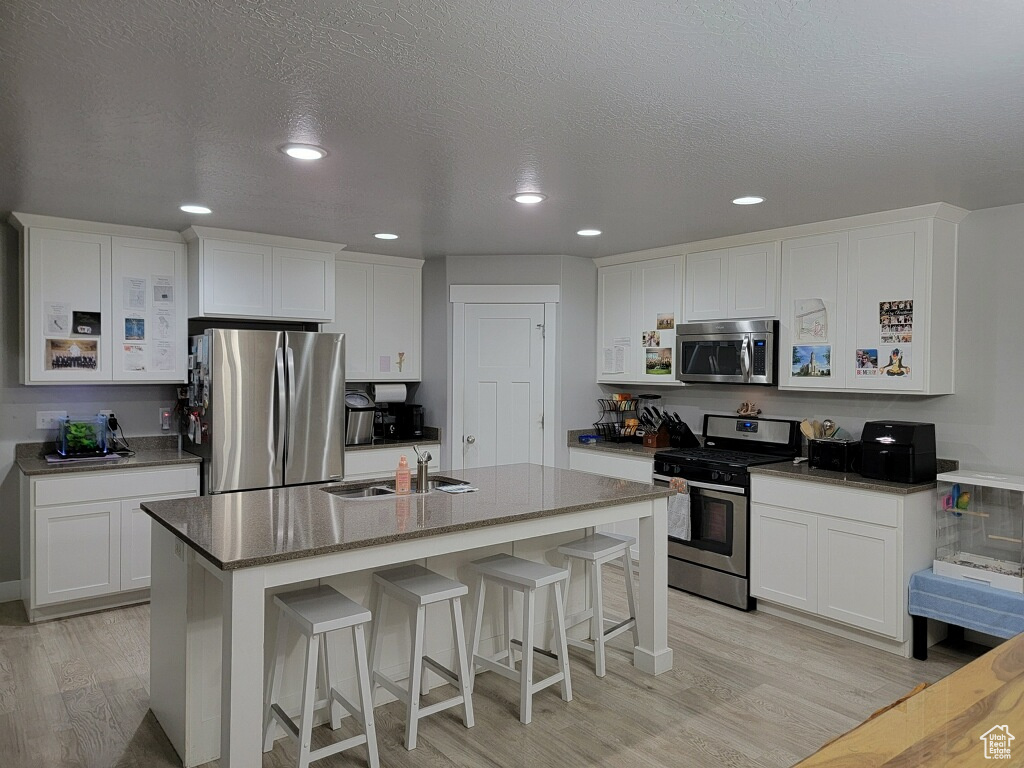 Kitchen with appliances with stainless steel finishes, a kitchen island with sink, white cabinetry, sink, and light wood-type flooring