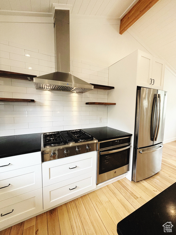 Kitchen featuring appliances with stainless steel finishes, light hardwood / wood-style flooring, lofted ceiling with beams, and wall chimney range hood