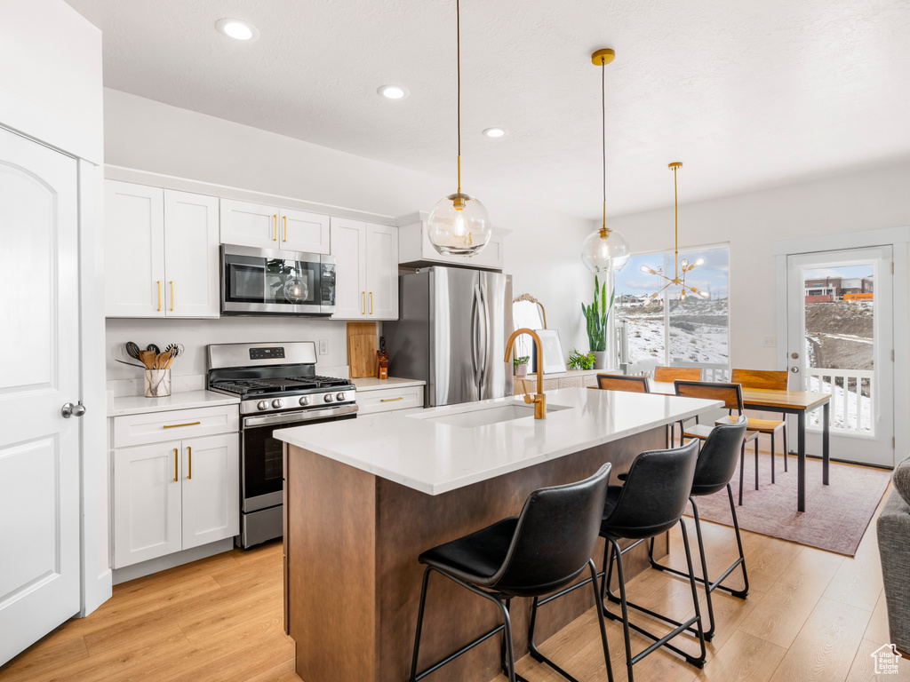 Kitchen featuring stainless steel appliances, a kitchen island with sink, sink, hanging light fixtures, and light wood-type flooring