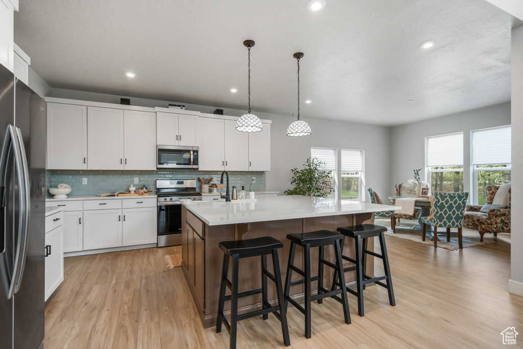 Kitchen featuring hanging light fixtures, white cabinets, light wood-type flooring, appliances with stainless steel finishes, and an island with sink