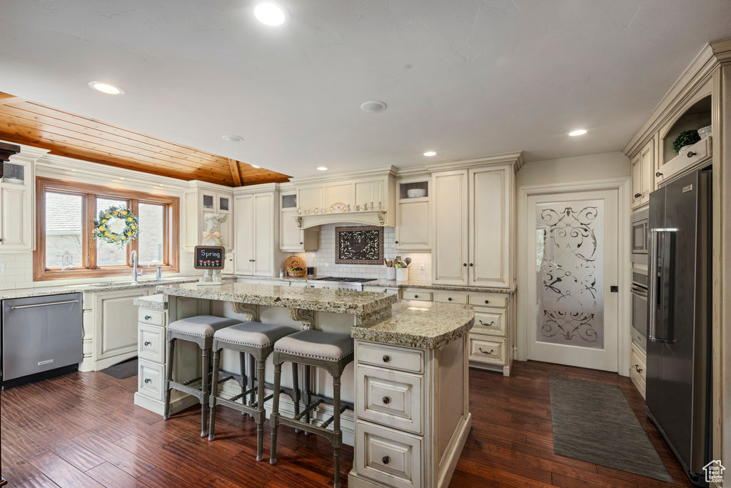 Kitchen with appliances with stainless steel finishes, a kitchen island, backsplash, dark wood-type flooring, and light stone countertops
