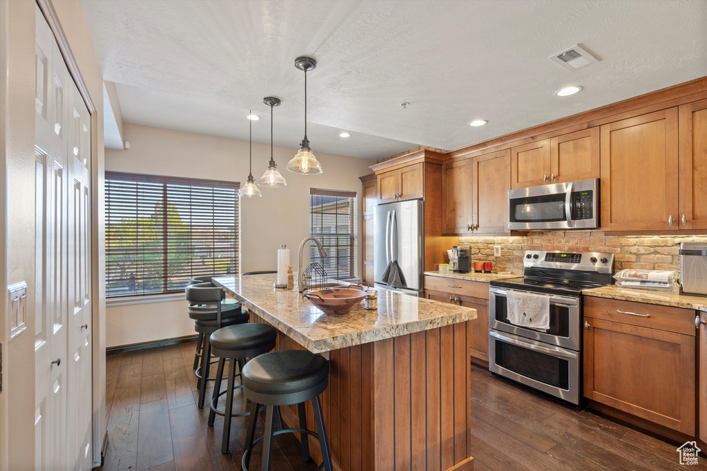 Kitchen with appliances with stainless steel finishes, dark hardwood / wood-style flooring, an island with sink, and hanging light fixtures
