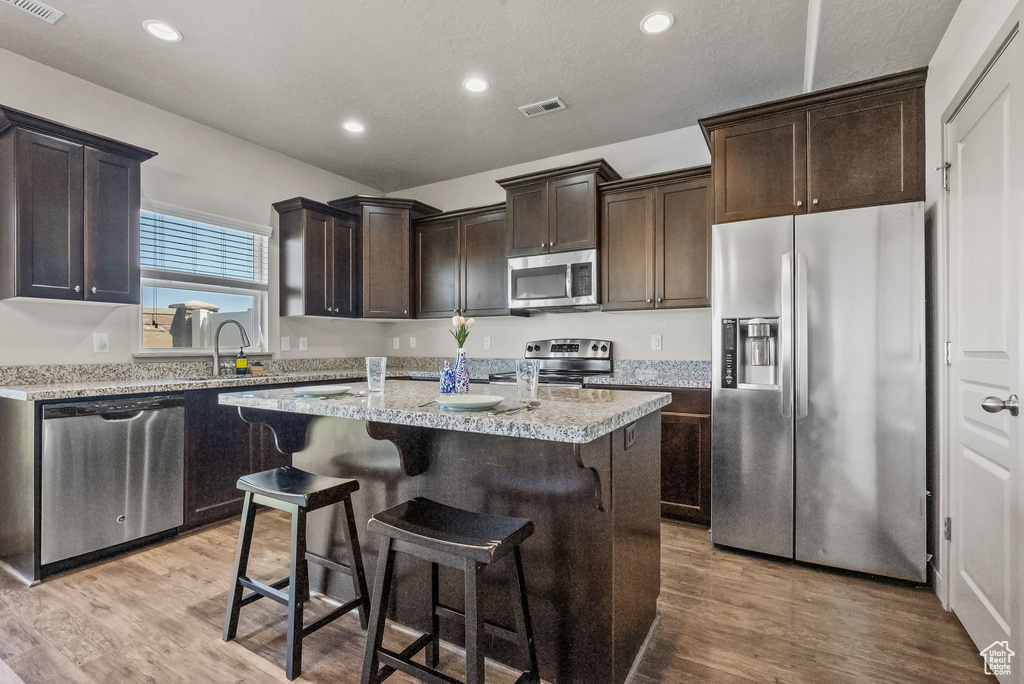 Kitchen featuring dark brown cabinetry, appliances with stainless steel finishes, a kitchen island, light hardwood / wood-style floors, and light stone countertops