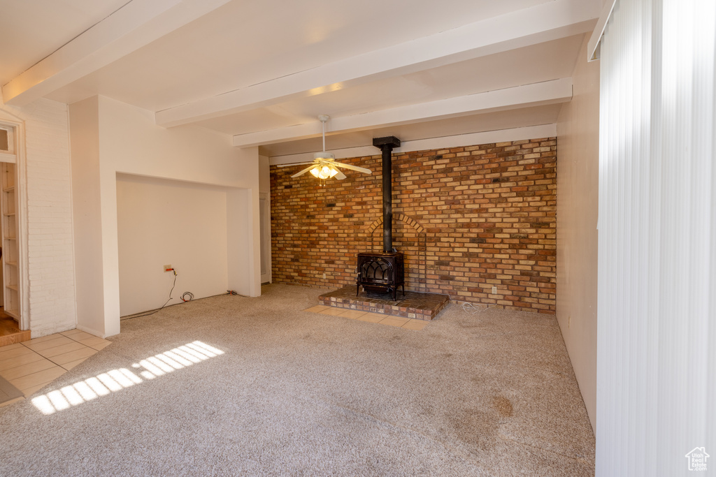 Unfurnished living room featuring carpet floors, ceiling fan, brick wall, beamed ceiling, and a wood stove
