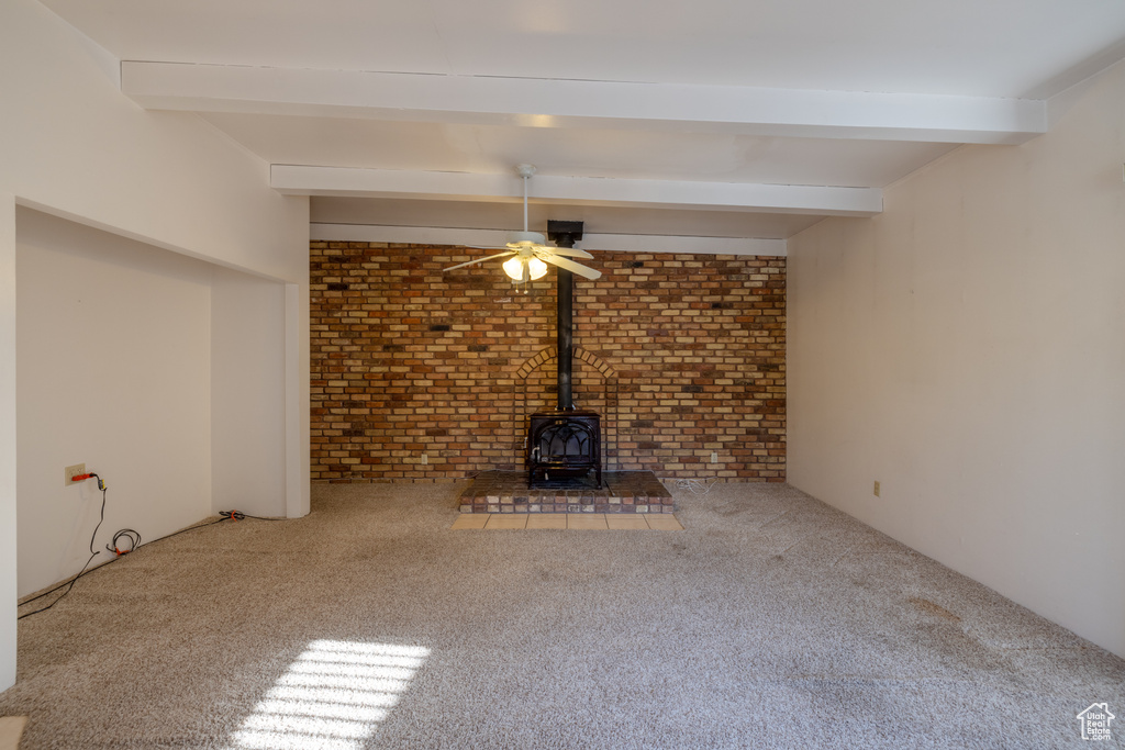 Unfurnished living room featuring ceiling fan, brick wall, beam ceiling, carpet flooring, and a wood stove