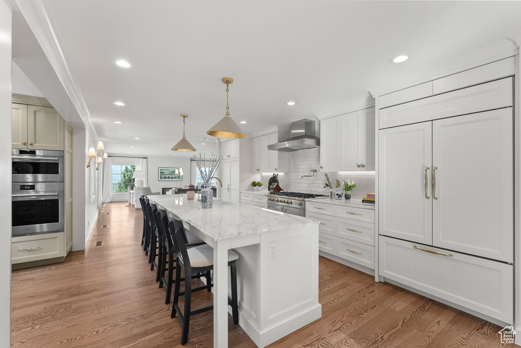 Kitchen featuring wall chimney exhaust hood, pendant lighting, light hardwood / wood-style flooring, appliances with stainless steel finishes, and an island with sink