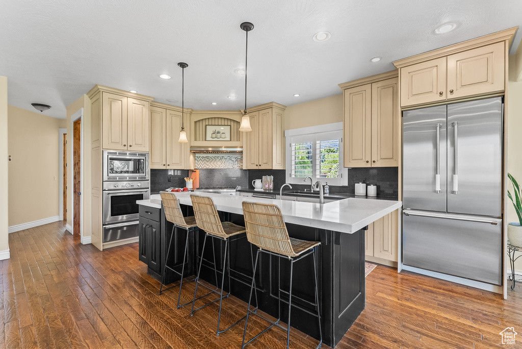 Kitchen with built in appliances, pendant lighting, hardwood / wood-style floors, a center island with sink, and tasteful backsplash