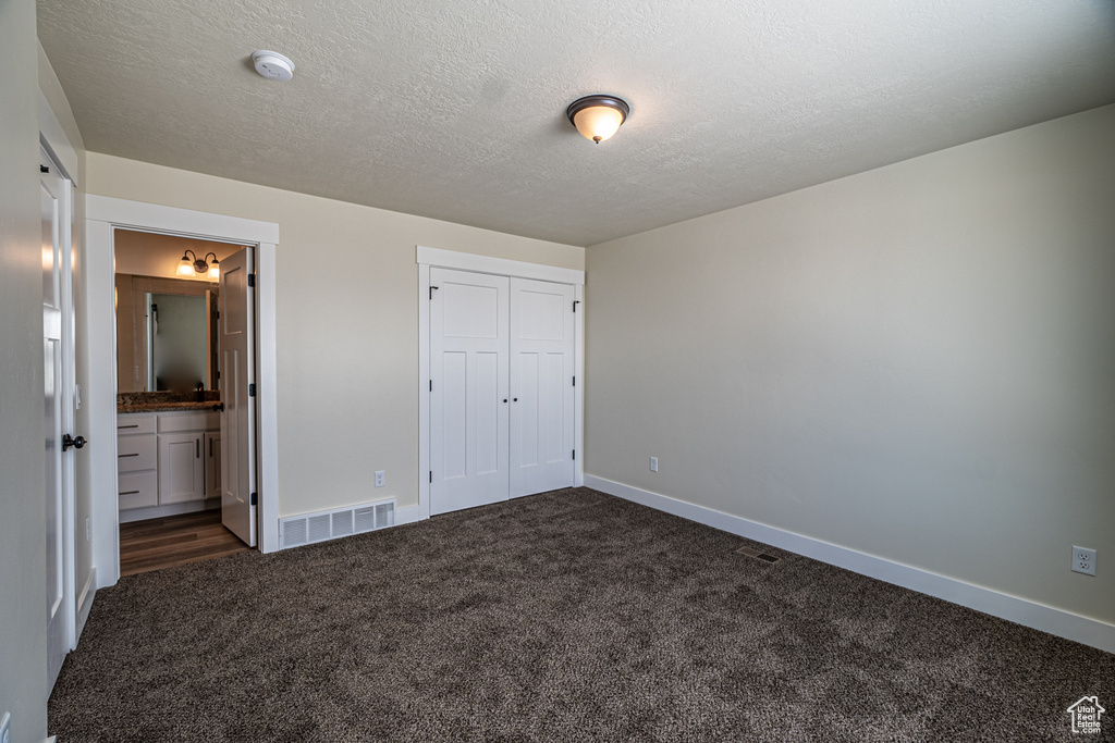 Unfurnished bedroom featuring a textured ceiling, a closet, ensuite bathroom, and dark colored carpet