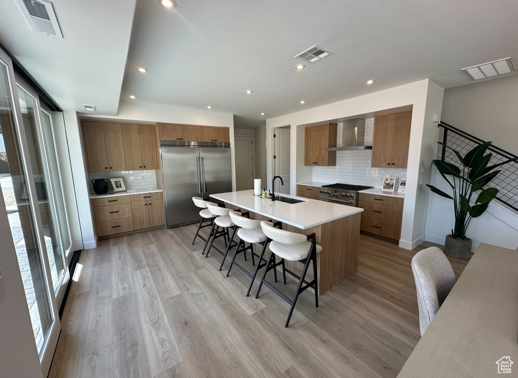 Kitchen featuring stainless steel built in refrigerator, a kitchen breakfast bar, light wood-type flooring, backsplash, and wall chimney range hood