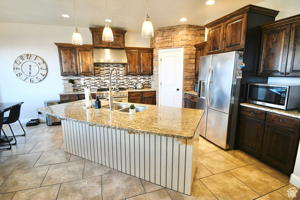Kitchen featuring wall chimney range hood, stainless steel appliances, light tile floors, tasteful backsplash, and an island with sink