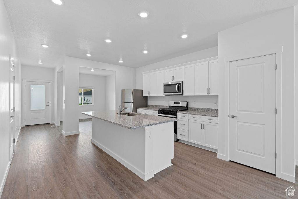 Kitchen with stainless steel appliances, a center island with sink, light stone counters, white cabinets, and hardwood / wood-style floors