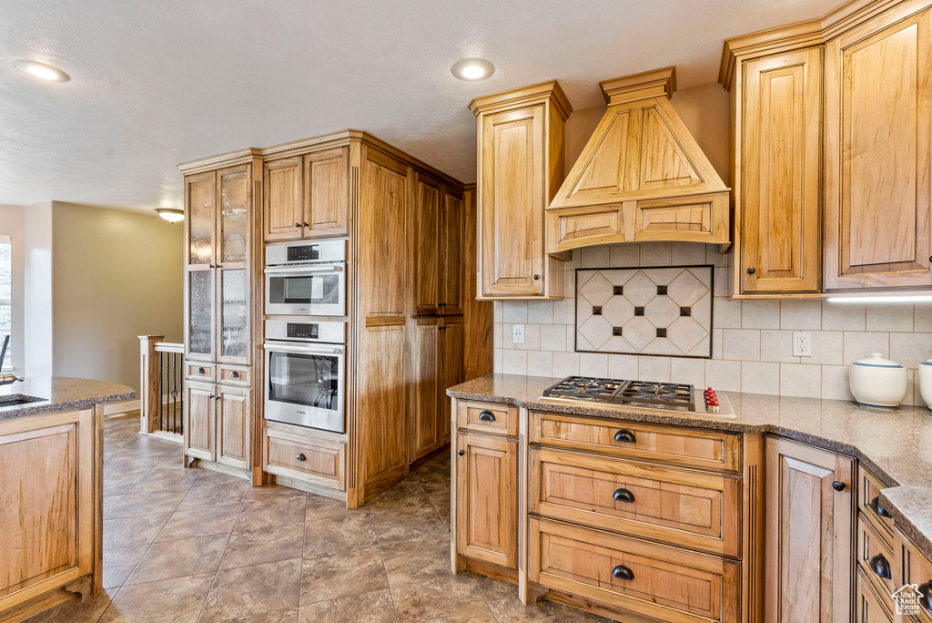 Kitchen with stainless steel double oven, backsplash, premium range hood, and light tile floors
