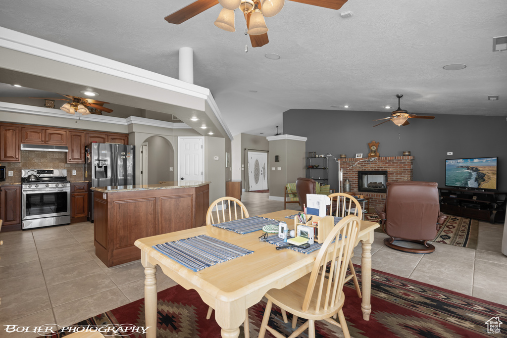 Tiled dining room with high vaulted ceiling, a brick fireplace, ceiling fan, and a textured ceiling