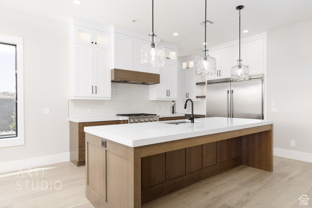 Kitchen featuring built in refrigerator, a center island with sink, light hardwood / wood-style floors, tasteful backsplash, and pendant lighting