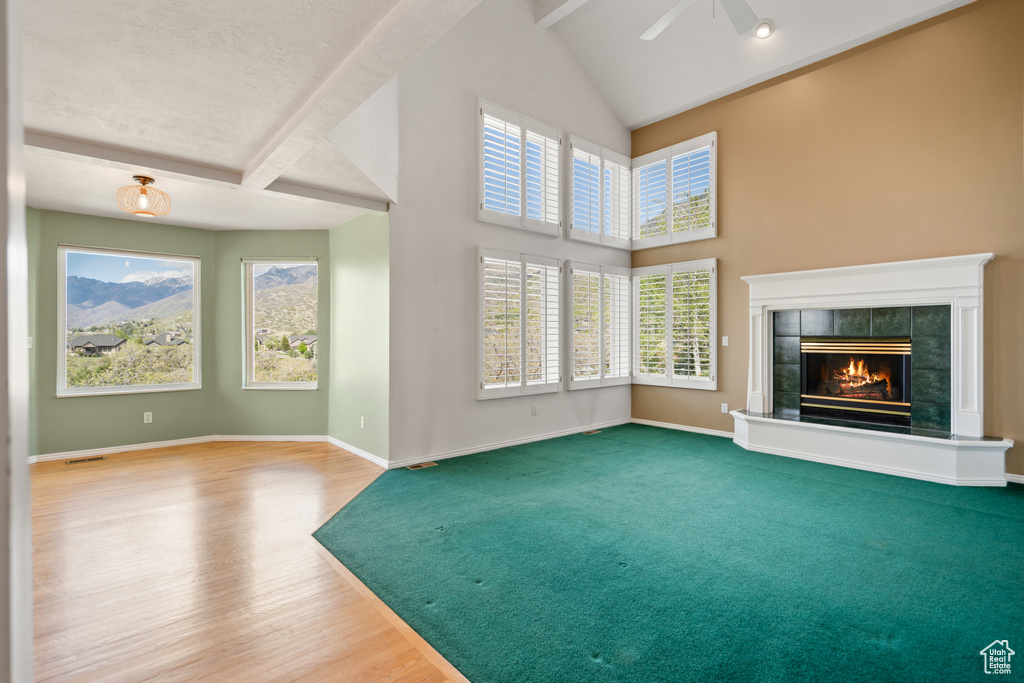 Unfurnished living room with high vaulted ceiling, carpet flooring, beam ceiling, a tile fireplace, and ceiling fan