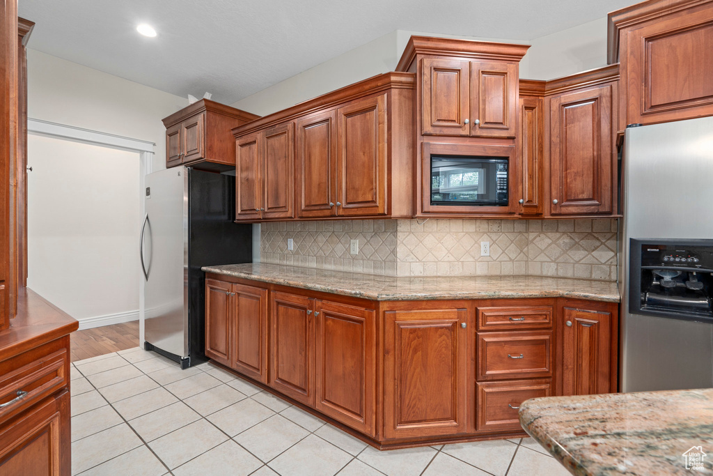 Kitchen featuring stainless steel fridge with ice dispenser, light tile flooring, tasteful backsplash, light stone counters, and black microwave