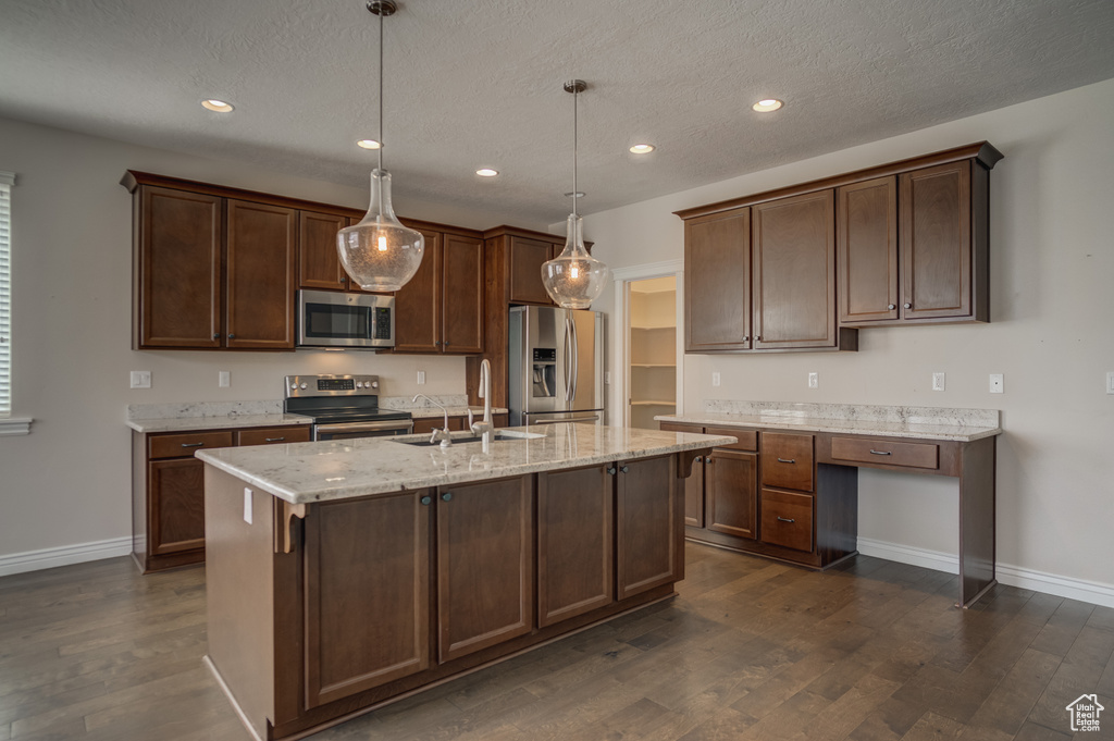 Kitchen with light stone counters, appliances with stainless steel finishes, dark wood-type flooring, hanging light fixtures, and a center island with sink