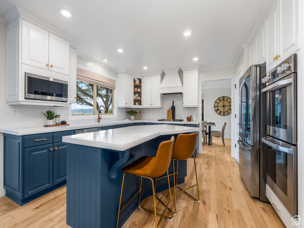 Kitchen featuring appliances with stainless steel finishes, white cabinetry, blue cabinetry, light hardwood / wood-style flooring, and custom range hood