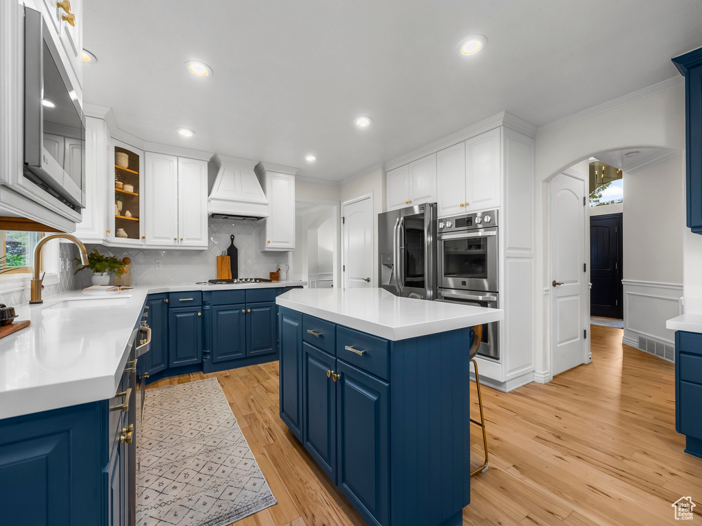 Kitchen with blue cabinets, appliances with stainless steel finishes, backsplash, light wood-type flooring, and custom range hood