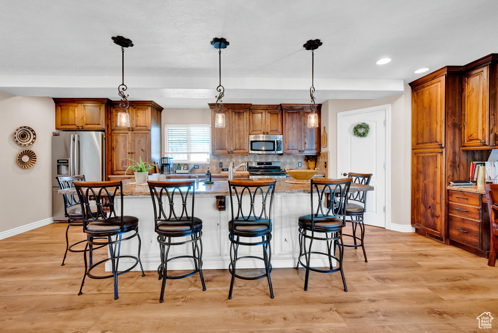 Kitchen with hanging light fixtures, light hardwood / wood-style flooring, appliances with stainless steel finishes, a center island with sink, and light stone counters