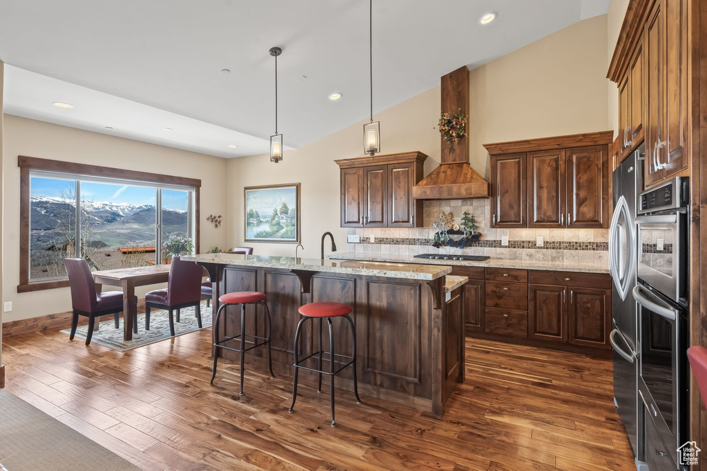 Kitchen with backsplash, dark wood-type flooring, hanging light fixtures, custom range hood, and a kitchen island with sink