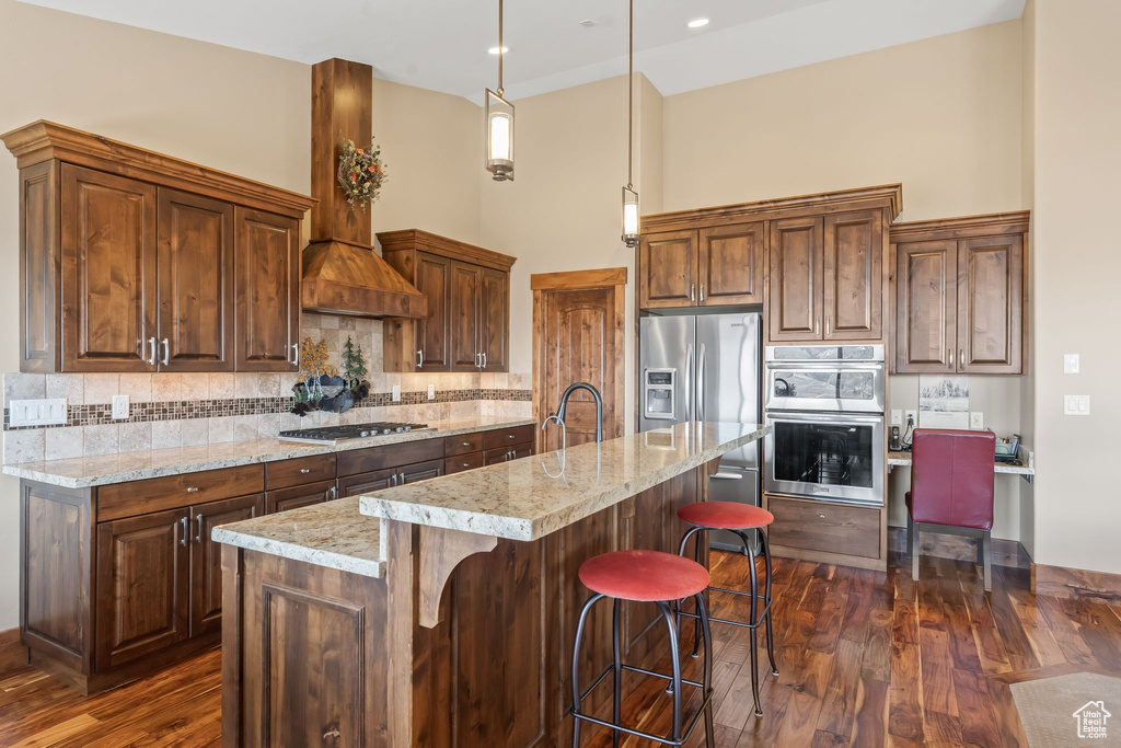Kitchen featuring hanging light fixtures, backsplash, a center island with sink, wall chimney range hood, and dark hardwood / wood-style flooring