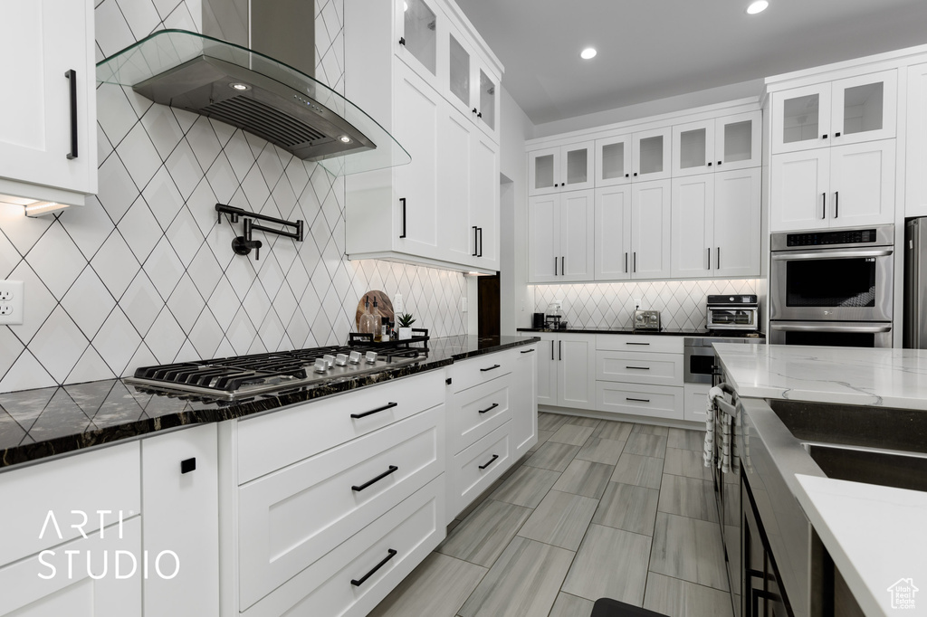 Kitchen featuring appliances with stainless steel finishes, tasteful backsplash, wall chimney range hood, white cabinetry, and dark stone countertops
