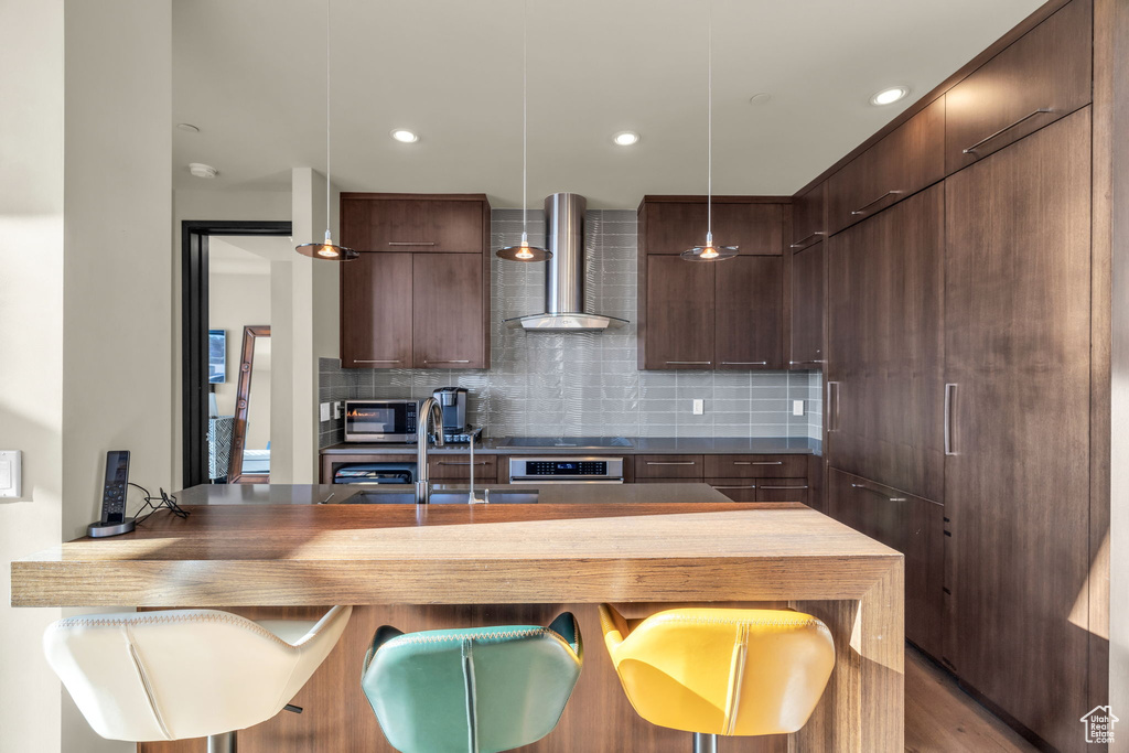 Kitchen with pendant lighting, wall chimney range hood, stainless steel appliances, hardwood / wood-style flooring, and tasteful backsplash