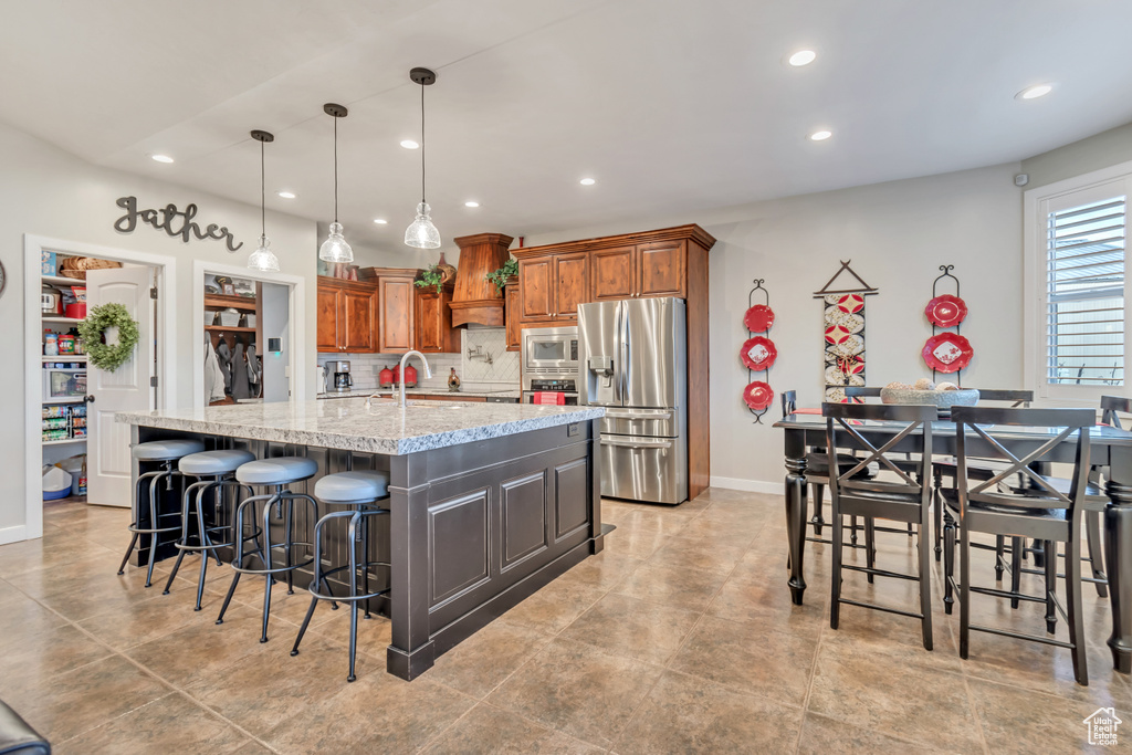 Kitchen featuring appliances with stainless steel finishes, light tile flooring, backsplash, an island with sink, and pendant lighting