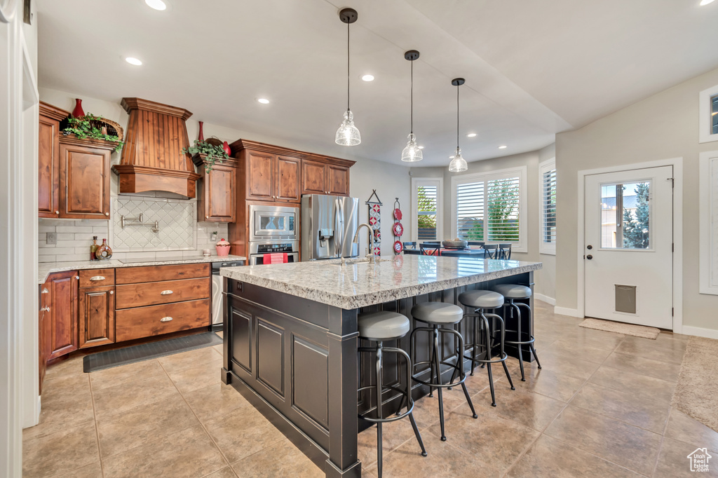 Kitchen with appliances with stainless steel finishes, pendant lighting, backsplash, custom range hood, and a kitchen island with sink