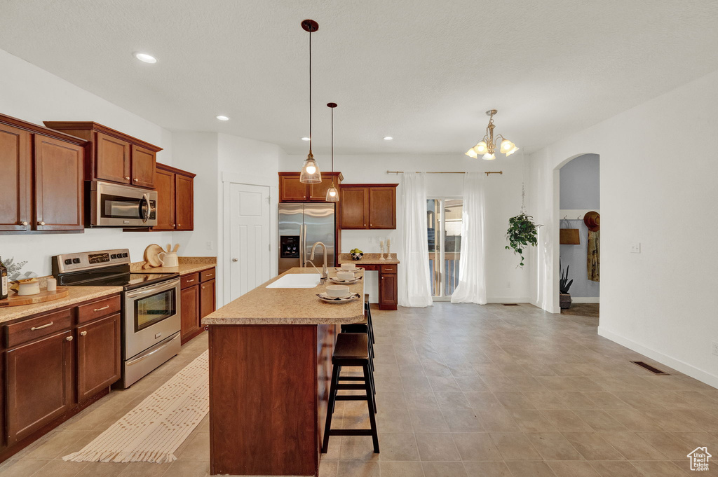 Kitchen with appliances with stainless steel finishes, hanging light fixtures, a breakfast bar area, light tile flooring, and an island with sink