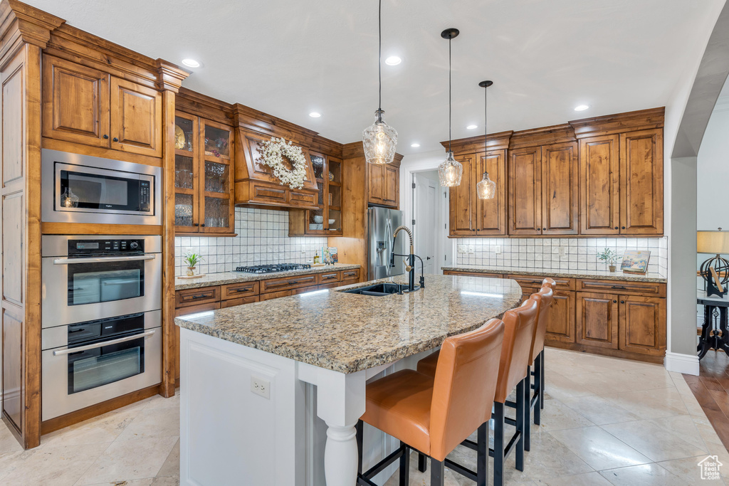 Kitchen with decorative light fixtures, appliances with stainless steel finishes, backsplash, custom range hood, and an island with sink
