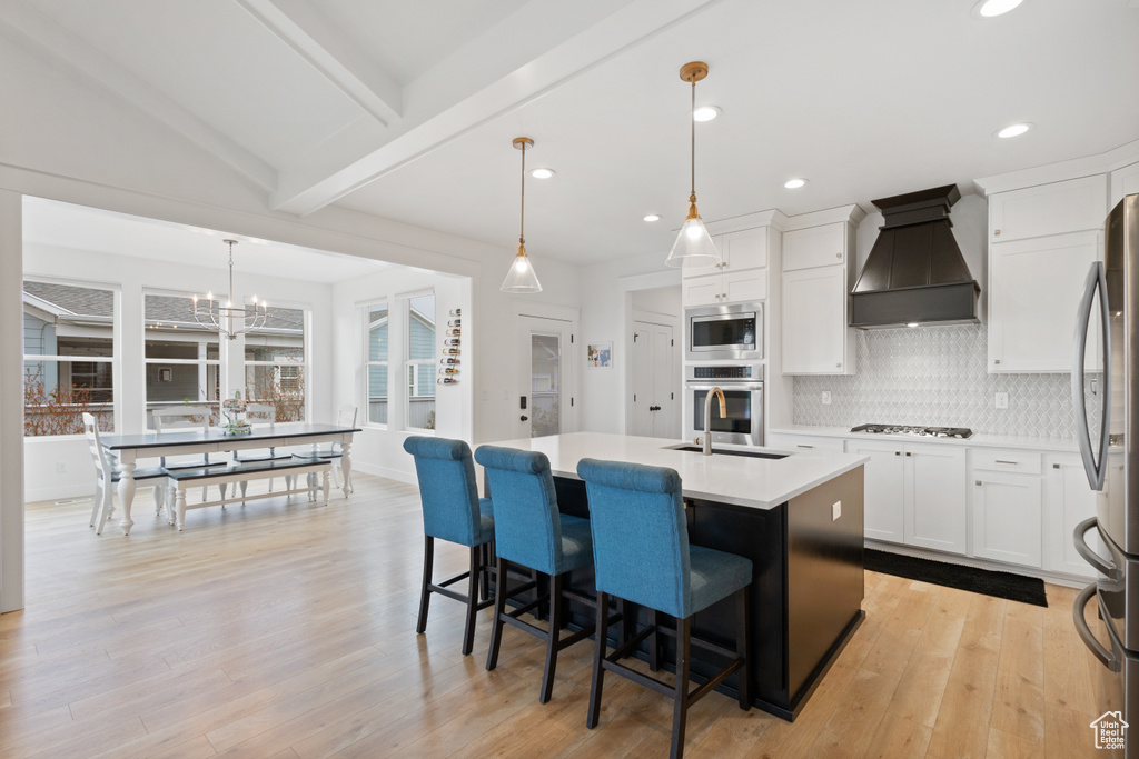 Kitchen featuring stainless steel appliances, light hardwood / wood-style floors, a center island with sink, lofted ceiling with beams, and custom exhaust hood