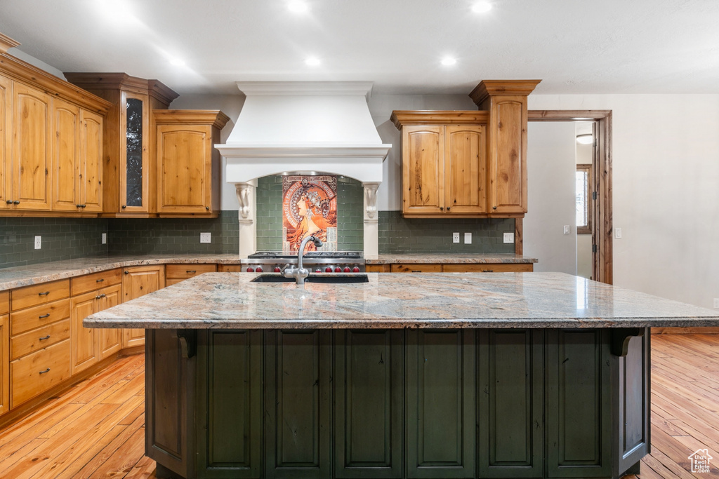 Kitchen featuring custom range hood, light stone countertops, backsplash, light hardwood / wood-style floors, and a kitchen island with sink