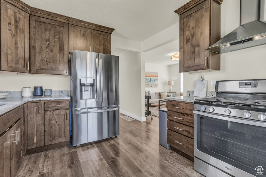 Kitchen featuring dark brown cabinets, light stone countertops, stainless steel appliances, wall chimney range hood, and dark hardwood / wood-style flooring