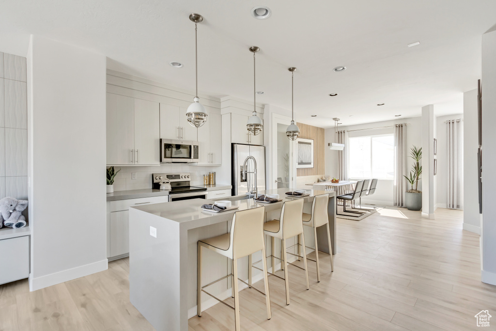 Kitchen featuring white cabinetry, appliances with stainless steel finishes, hanging light fixtures, light hardwood / wood-style flooring, and a kitchen island with sink