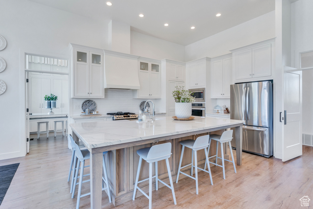 Kitchen with appliances with stainless steel finishes, a center island with sink, custom exhaust hood, white cabinets, and light wood-type flooring